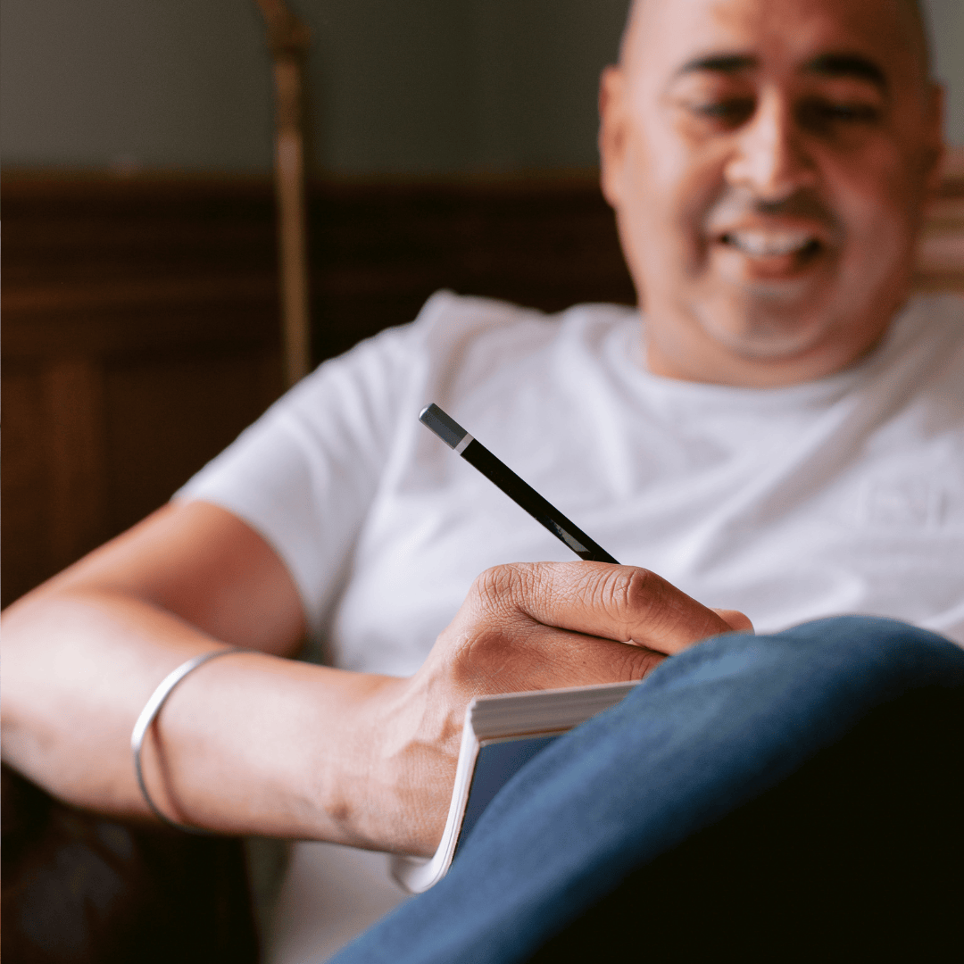 Man sitting on a couch, writing in a notebook with a pen.
