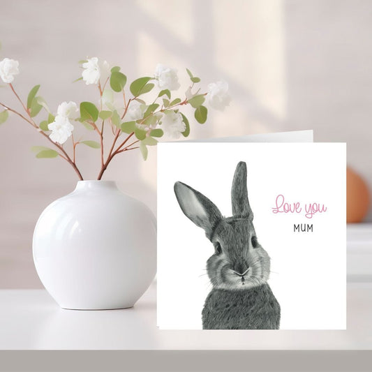A hand-drawn 'Love You Mum' bunny greeting card, placed next to a vase of flowers. Perfect for Mother's Day, birthdays, or just to show appreciation, this card features an adorable bunny design. Printed on high-quality cardstock, the interior is blank, ready for a heartfelt message.