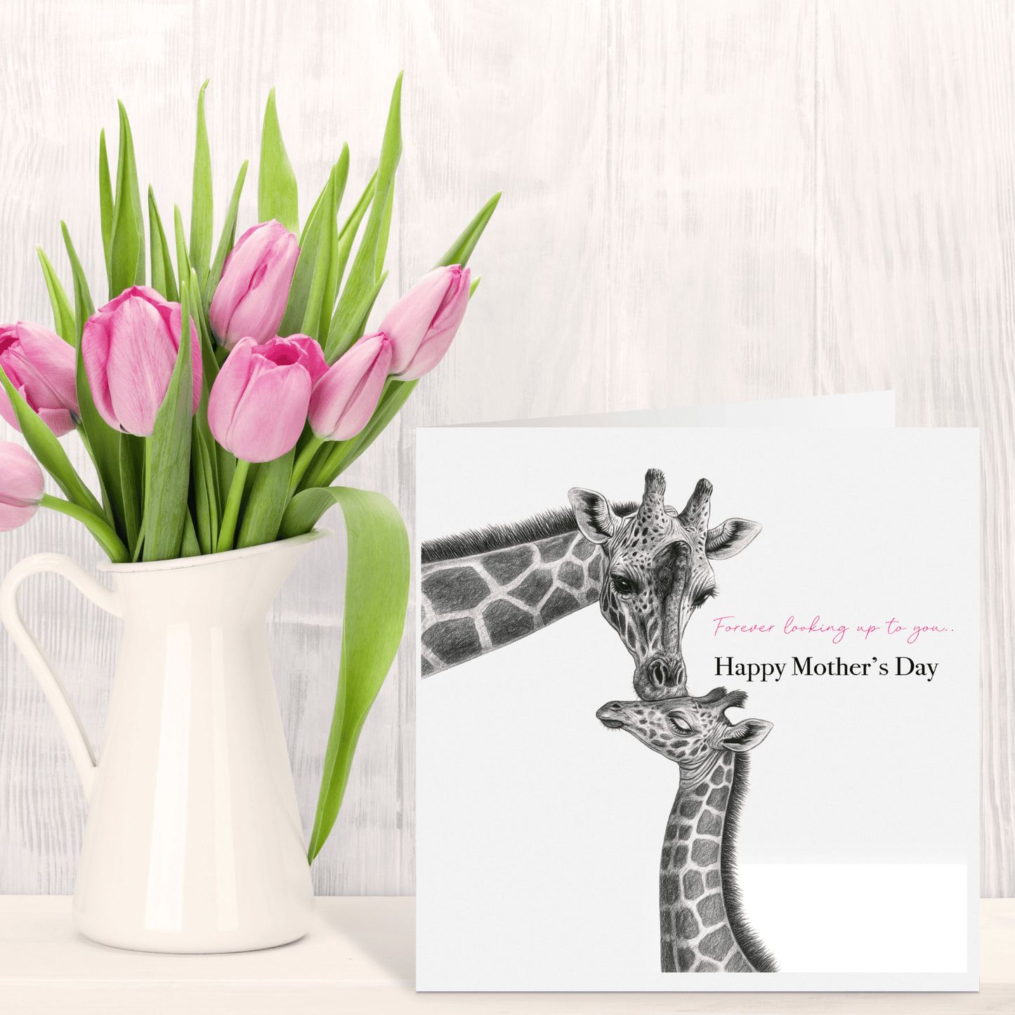 Pink tulips in a white pitcher next to a 'Happy Mother's Day' card with giraffes on a light wooden surface.