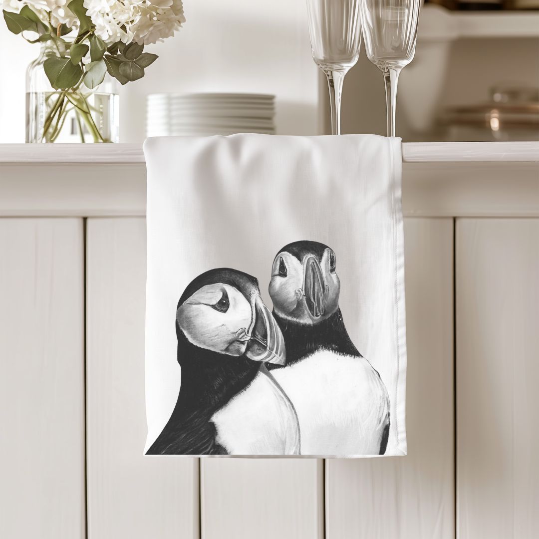 A premium cotton tea towel featuring a hand-drawn illustration of two puffins, adding a whimsical touch to your kitchen decor. Made from soft, absorbent cotton, it's both practical and charming. Ideal for bird enthusiasts and coastal art lovers.
