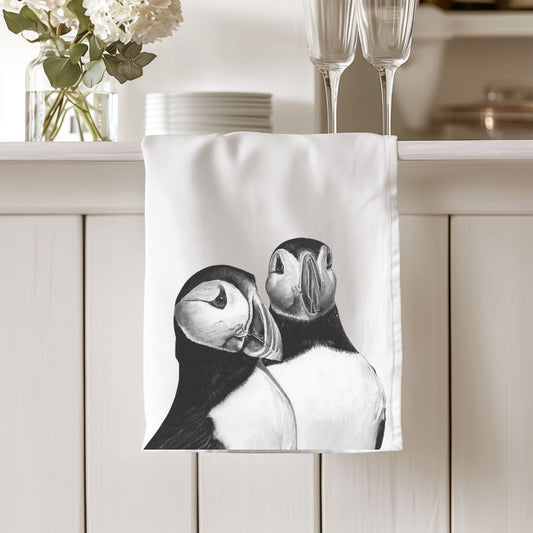 A premium cotton tea towel featuring a hand-drawn illustration of two puffins, adding a whimsical touch to your kitchen decor. Made from soft, absorbent cotton, it's both practical and charming. Ideal for bird enthusiasts and coastal art lovers.