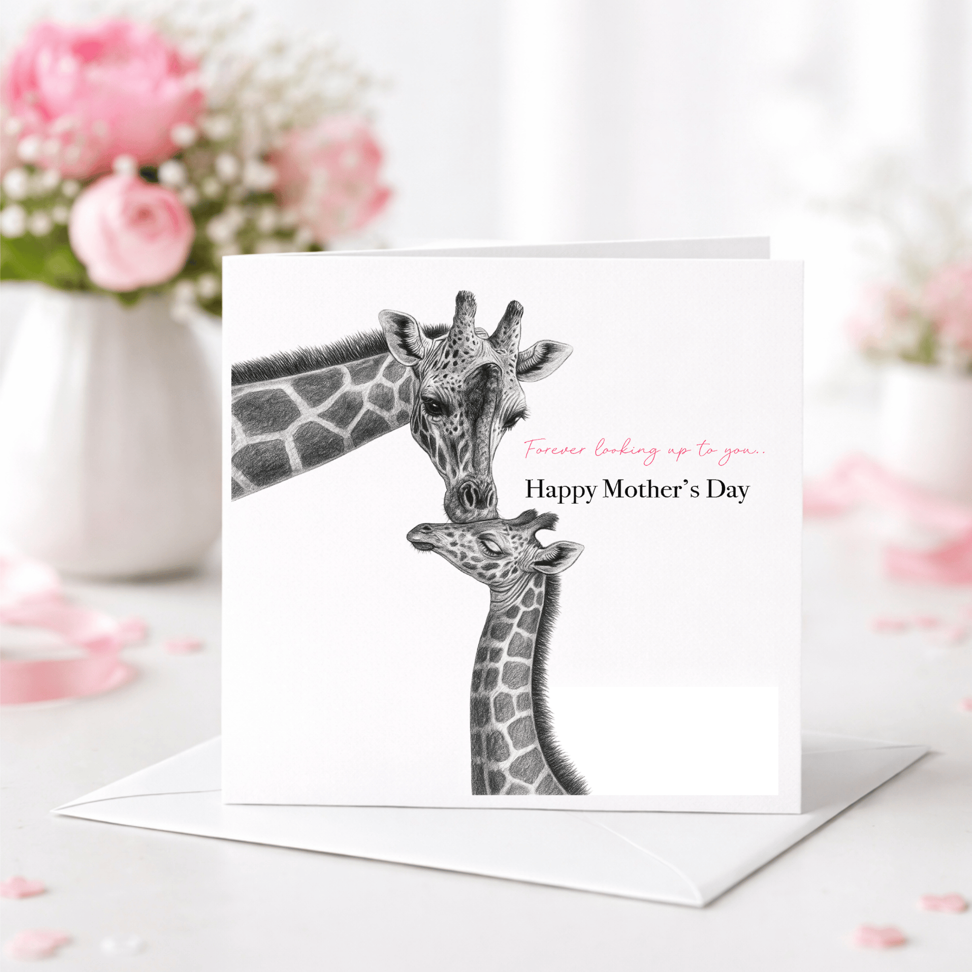 Greeting card with illustration of two giraffes and 'Happy Mother's Day' text, placed on a surface with flowers in the background.