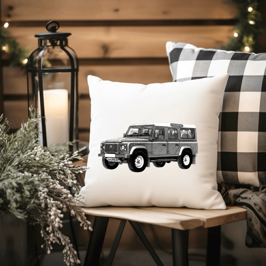 White pillow with a black and white illustration of a car on a wooden bench with a candle and plaid blanket in the background.