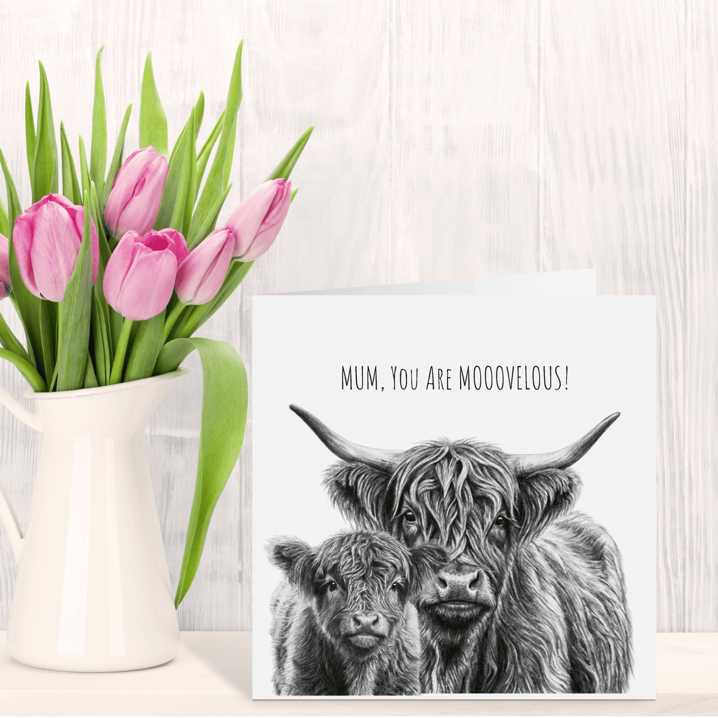 Pink tulips in a white vase next to a card with Highland cow illustration and text on a wooden surface.