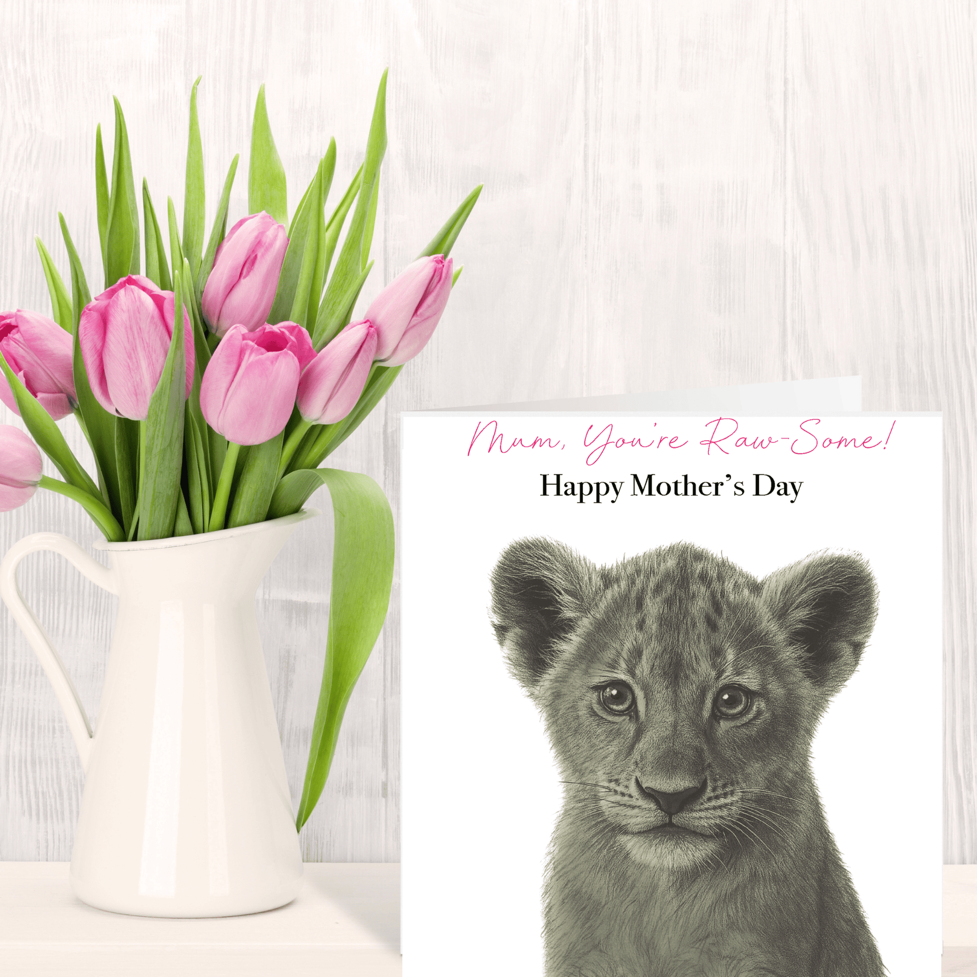 Pink tulips in a white vase next to a card with a lion cub and 'Happy Mother's Day' text on a light wooden background.