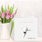 Pink tulips in a white vase next to a card with an airplane and text on a light wooden surface.