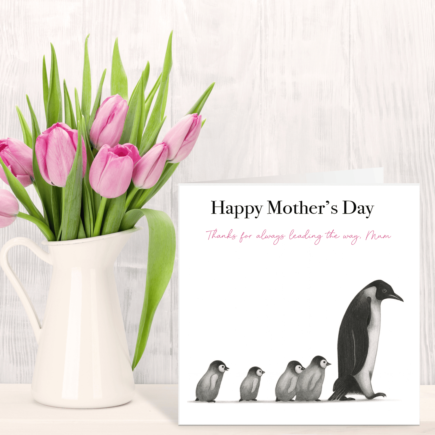Pink tulips in a white pitcher next to a Mother's Day card with penguin design on a light wooden surface.
