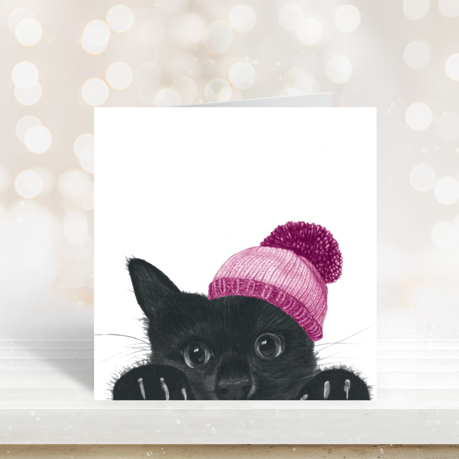 “Hand-drawn winter greeting card featuring a cute cat peeking out wearing a pink bobble hat.