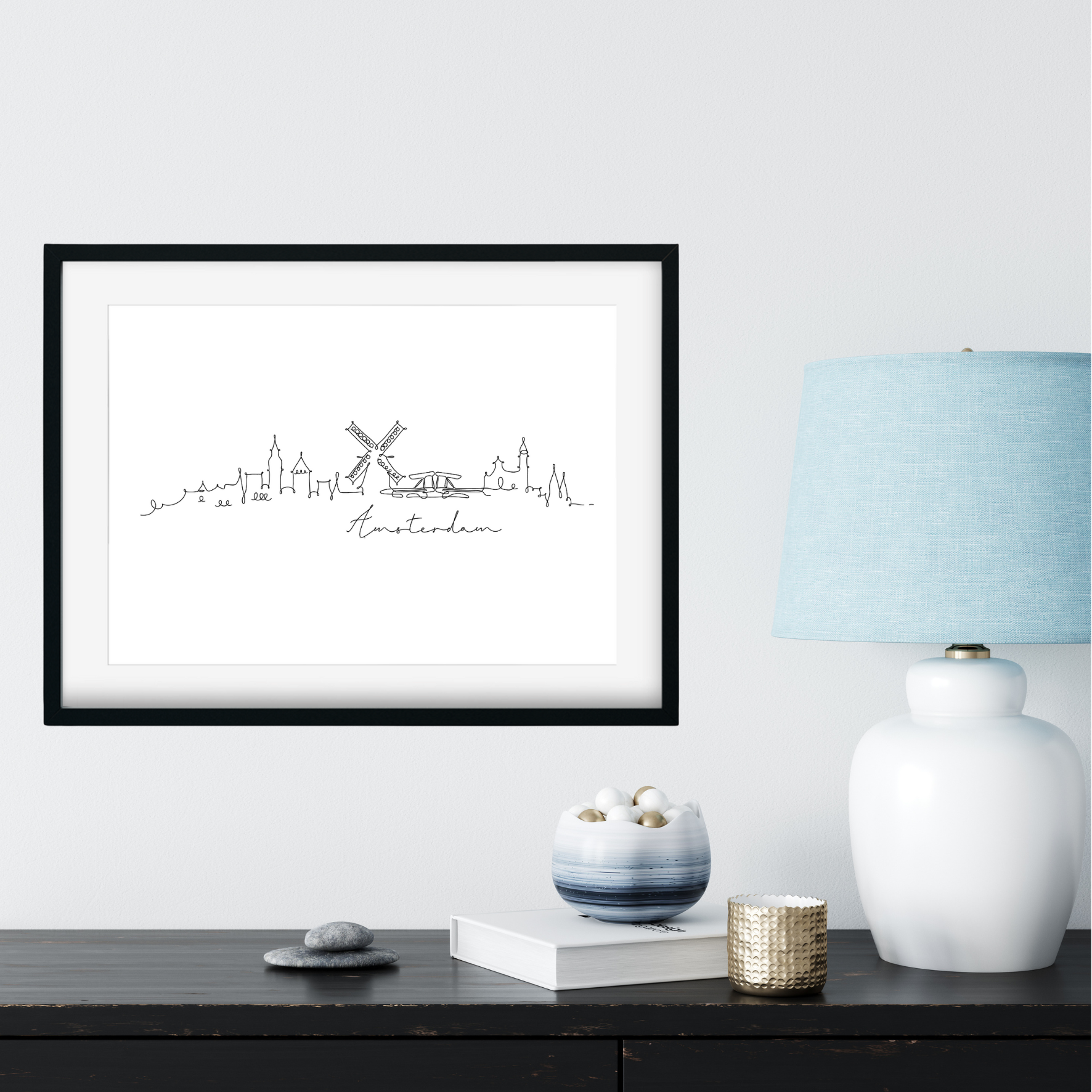 Framed artwork of a city skyline with a windmill on a white wall above a black shelf.