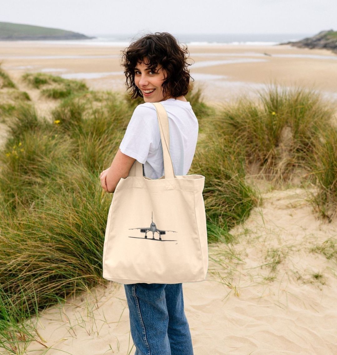 A woman using the large organic Concorde shopper bag at the beach, showcasing its spacious design and eco-friendly canvas material. The bag features a hand-drawn illustration of the iconic Concorde aircraft, adding a stylish touch to this functional accessory. Perfect for carrying beach essentials like towels, sunscreen, and snacks, this durable and sustainable bag is ideal for both aviation lovers and those seeking a chic, practical beach com