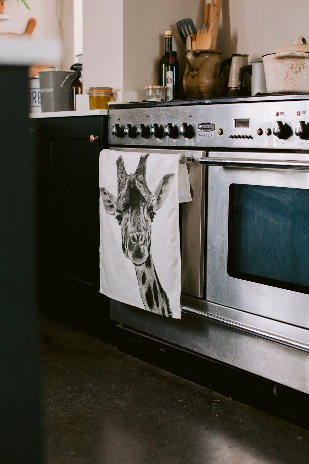 A hand-drawn giraffe tea towel, made from premium cotton, hanging over an oven door. With its unique wildlife-inspired design, this towel adds a whimsical touch to your kitchen. A perfect gift for animal lovers, it combines practicality with a delightful nature theme, making it a lovely addition to any home.